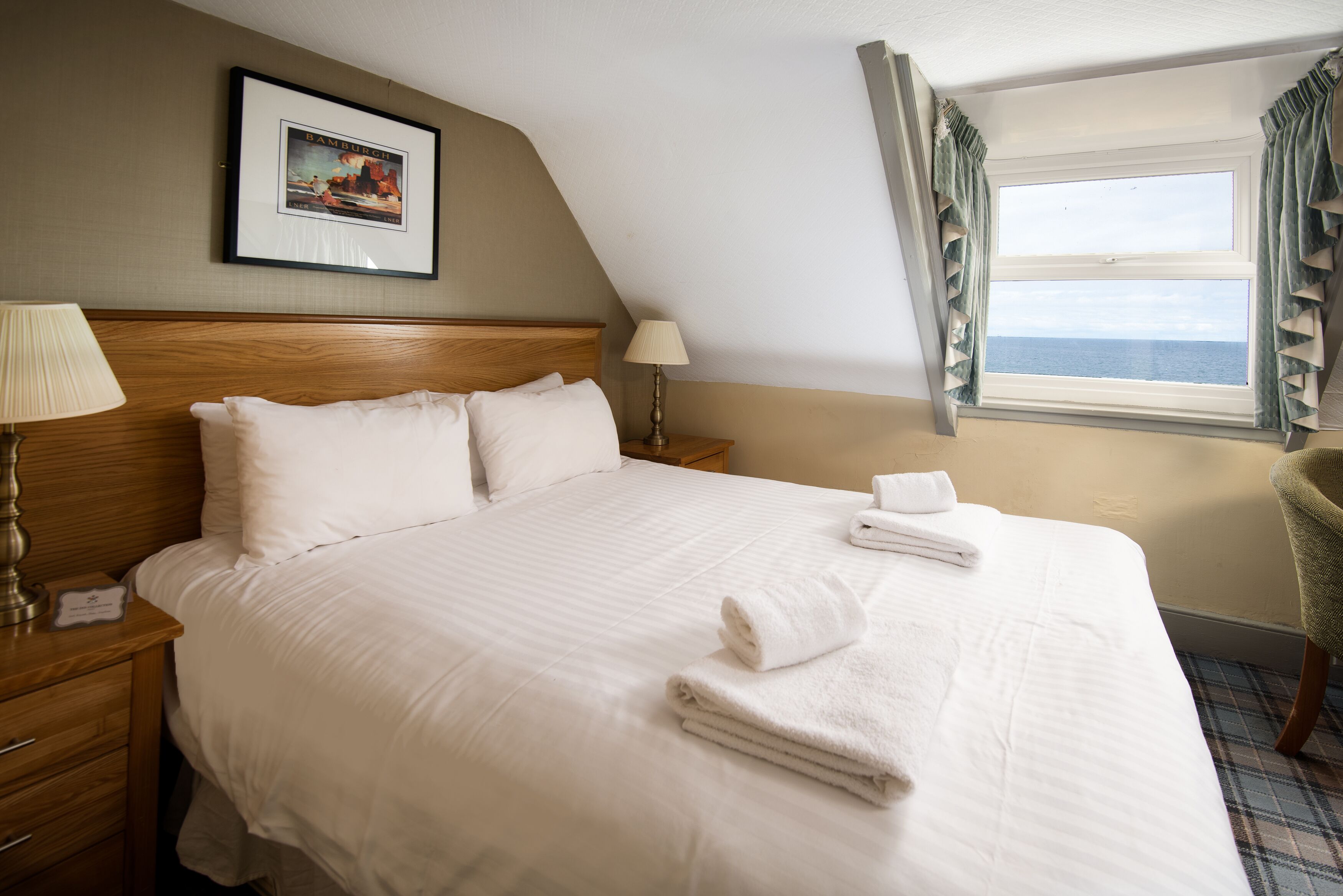 A neatly made bed with white linens and towels, a wooden headboard, a framed picture on the wall, and a window with curtains.