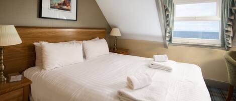 A neatly made bed with white linens and towels, a wooden headboard, a framed picture on the wall, and a window with curtains.