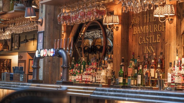 Een barruimte met houten inrichting, planken met flessen en een bordje met de tekst 'The Bamburgh Castle Inn'.