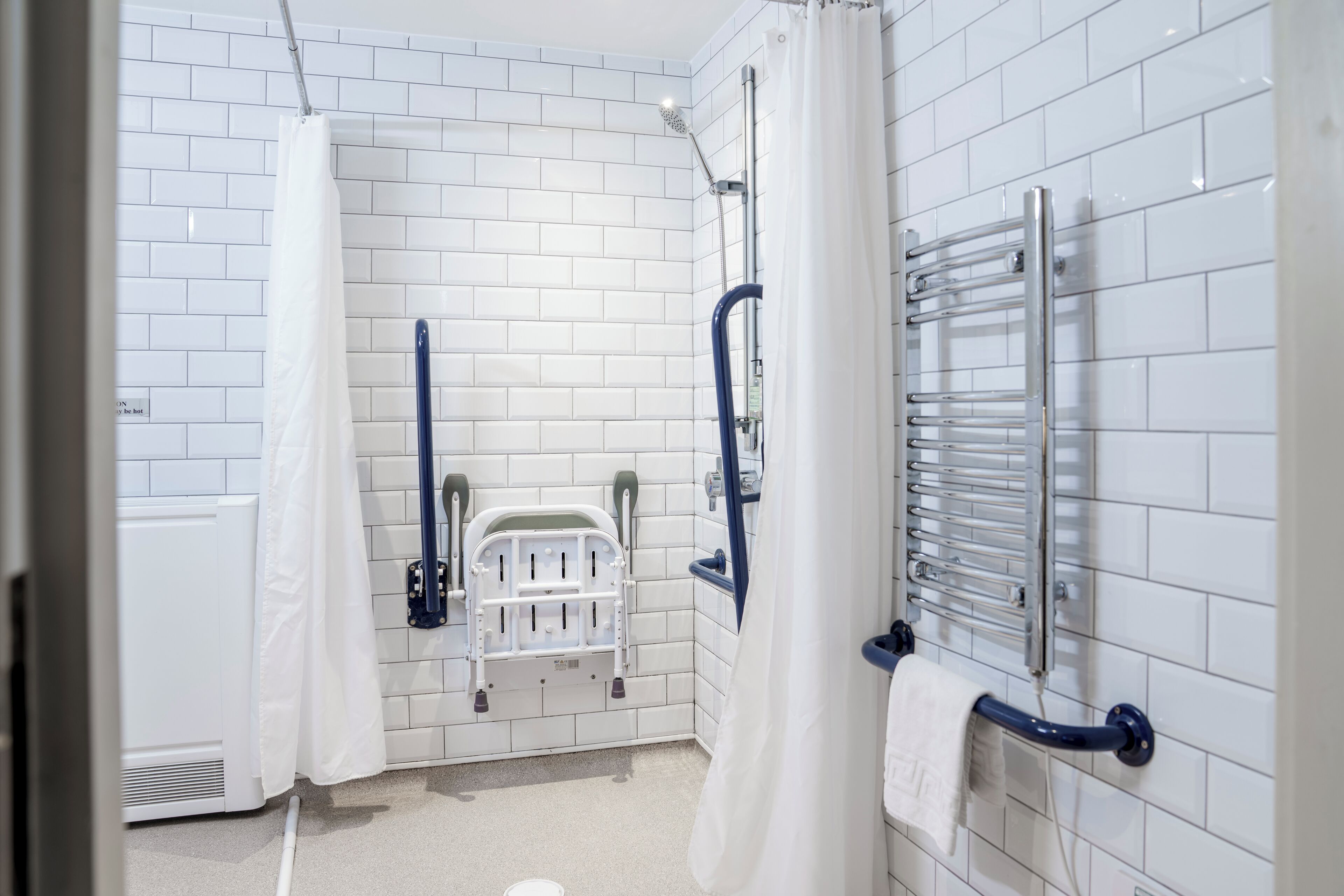 A bathroom with white tiled walls, a white shower curtain, a white toilet, a blue grab bar, and a white towel rack.