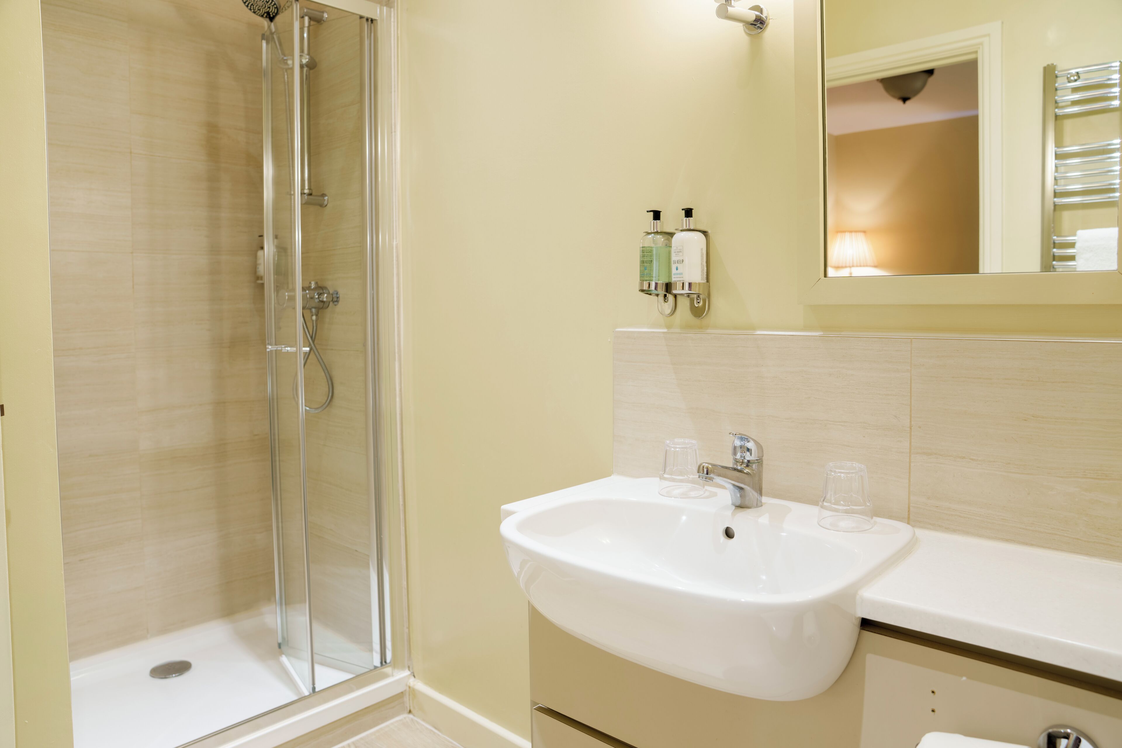 A bathroom with a glass shower enclosure, a white sink with two glasses, and toiletries on the wall.