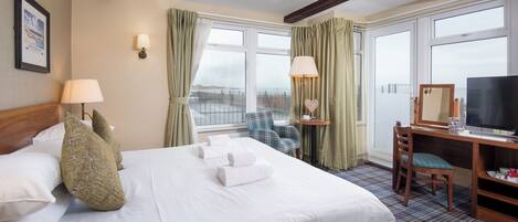 A hotel room with a large bed, a desk with a chair, a television, and a view of the city through the windows.