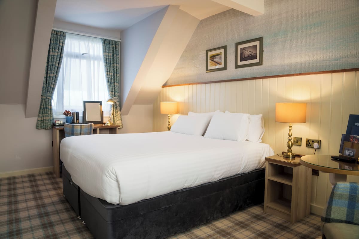Ein Hotelzimmer mit einem großen Bett, zwei Nachttischlampen, einem kleinen Tisch, einem Stuhl und einem Fenster mit Vorhängen.