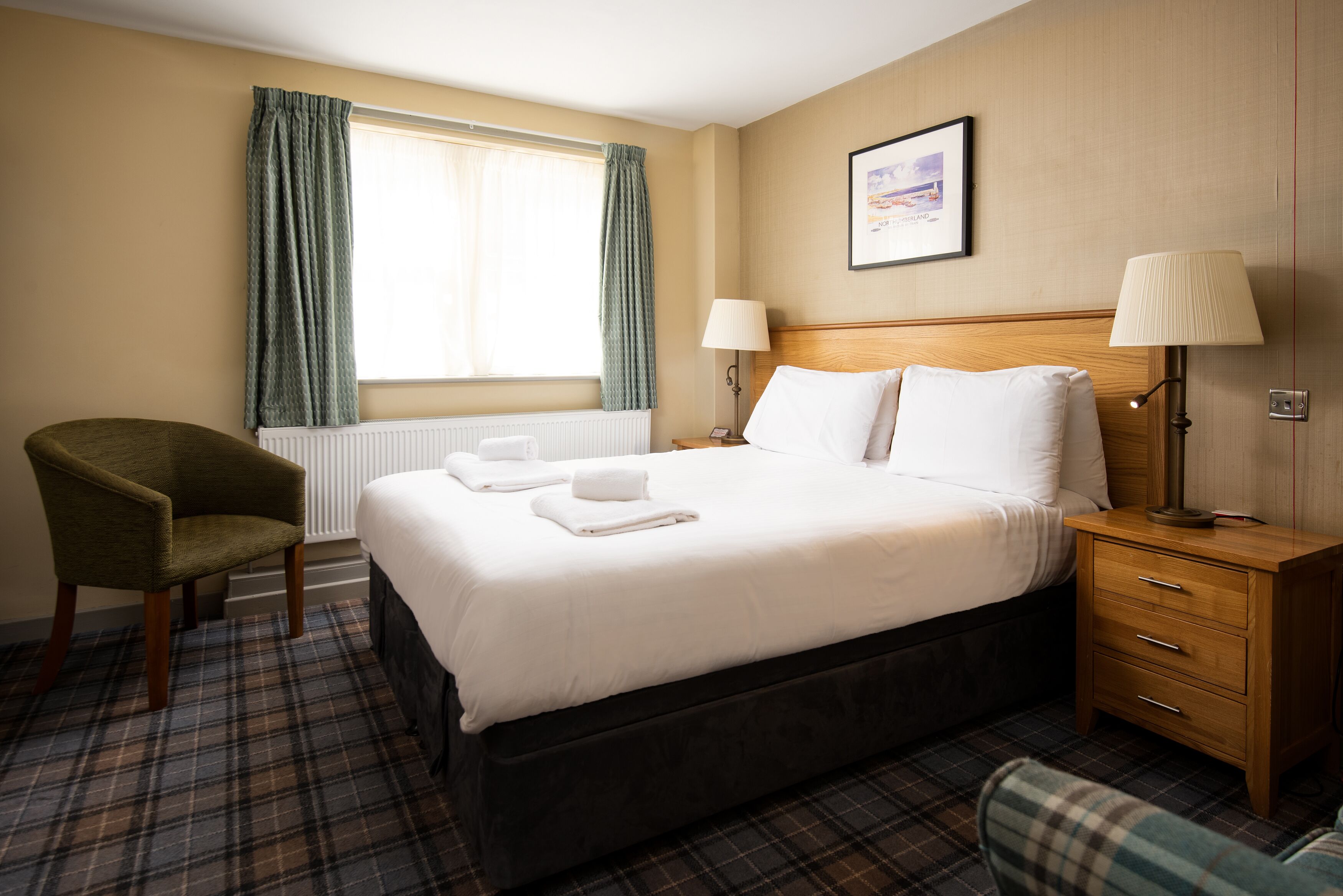 A hotel room with a bed, a chair, a nightstand, a lamp, and a window with curtains.