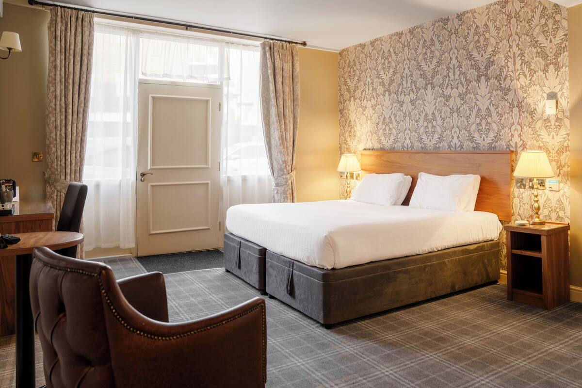 Ein Hotelzimmer mit einem großen Bett, zwei Nachttischen mit Lampen, einem Sessel, einem Schreibtisch und einer Tür.