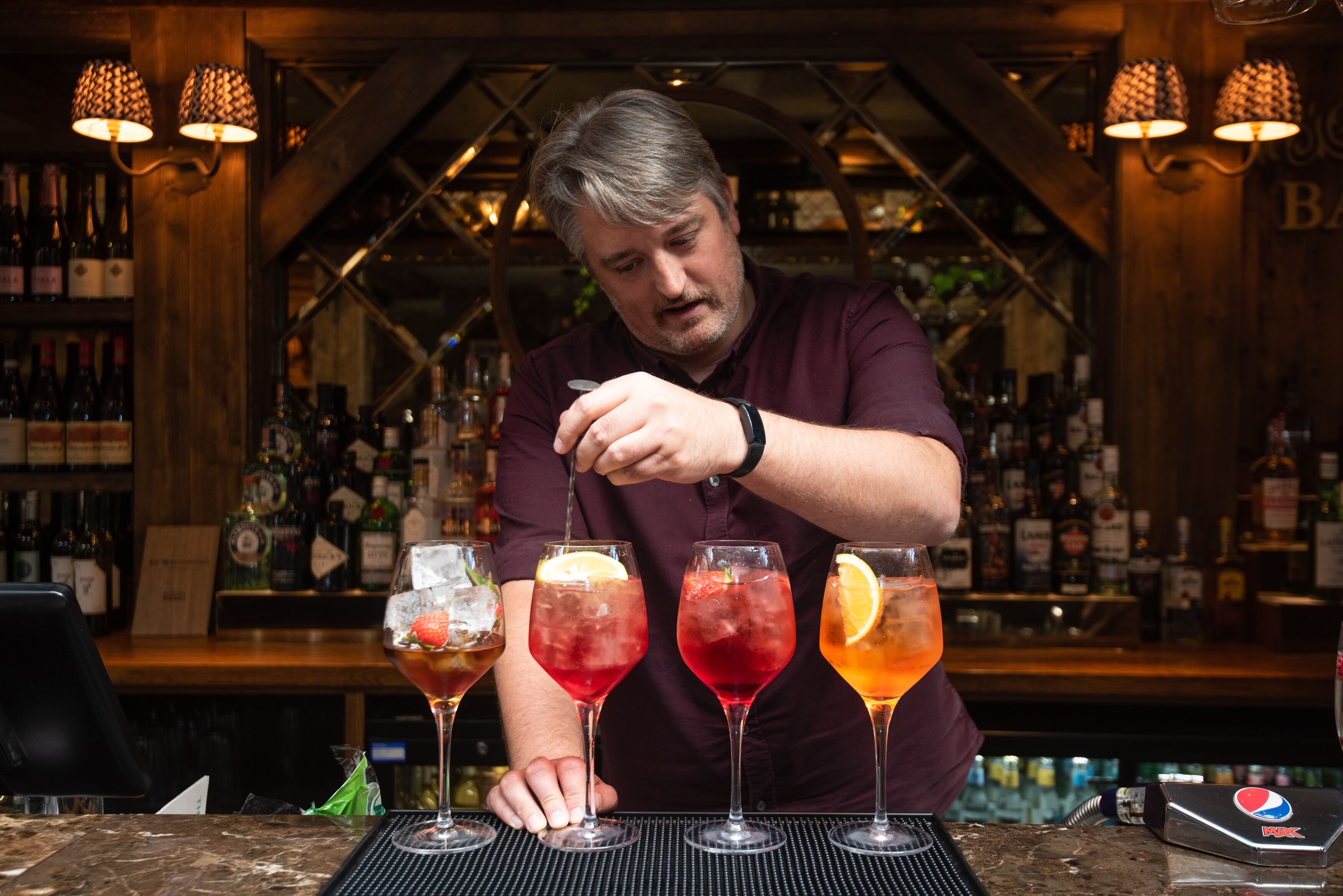 A bartender preparing drinks at a bar with a wooden interior and a variety of bottles in the background.