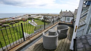 A balcony with wicker furniture, a view of the sea, and a parking area with cars.