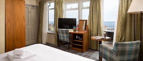 A hotel room with a bed, a television, a desk with a chair, and a view of the sea.