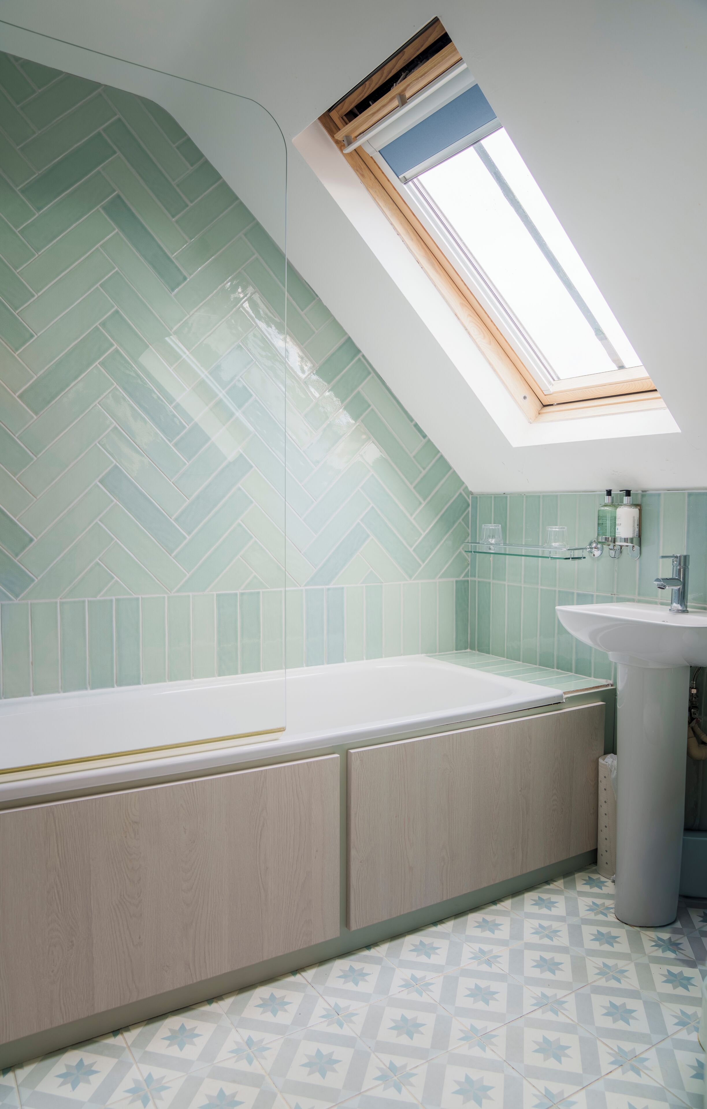 A modern bathroom with a herringbone tile backsplash, a skylight, a freestanding bathtub, and a pedestal sink.