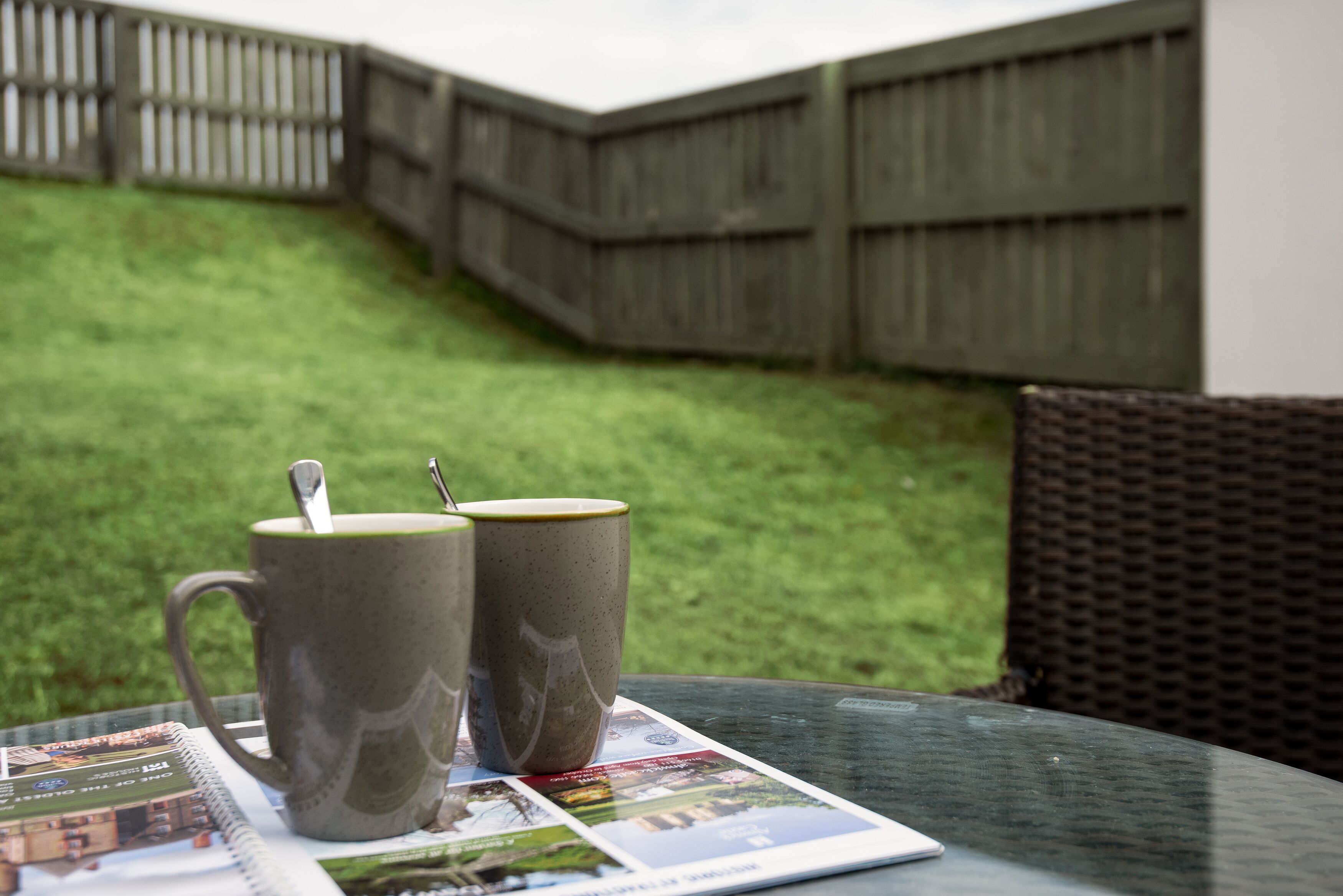A table with two mugs and a spoon, a magazine, and a booklet on a glass table outdoors.