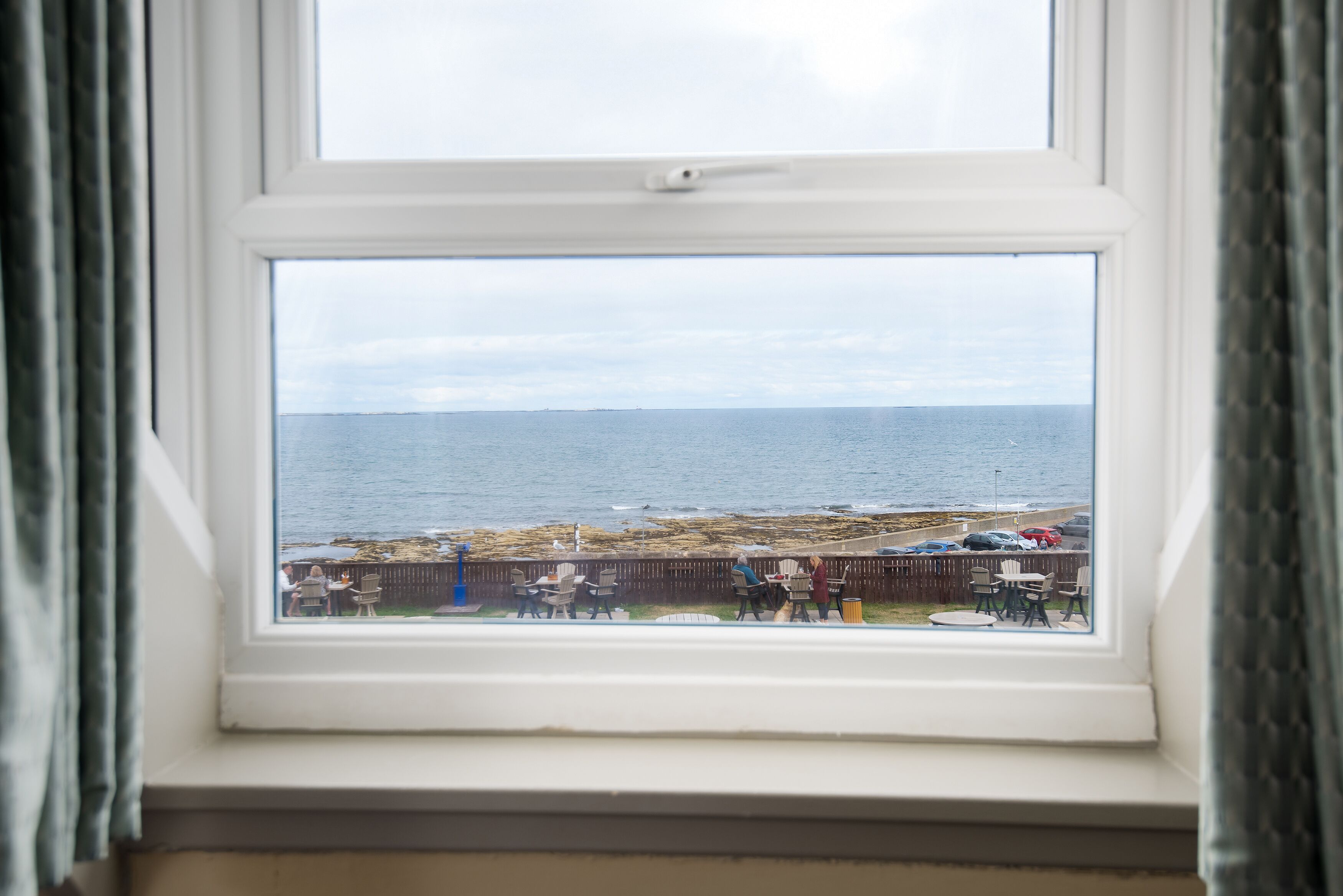 A window with a view of the sea and a beach area with people and chairs.