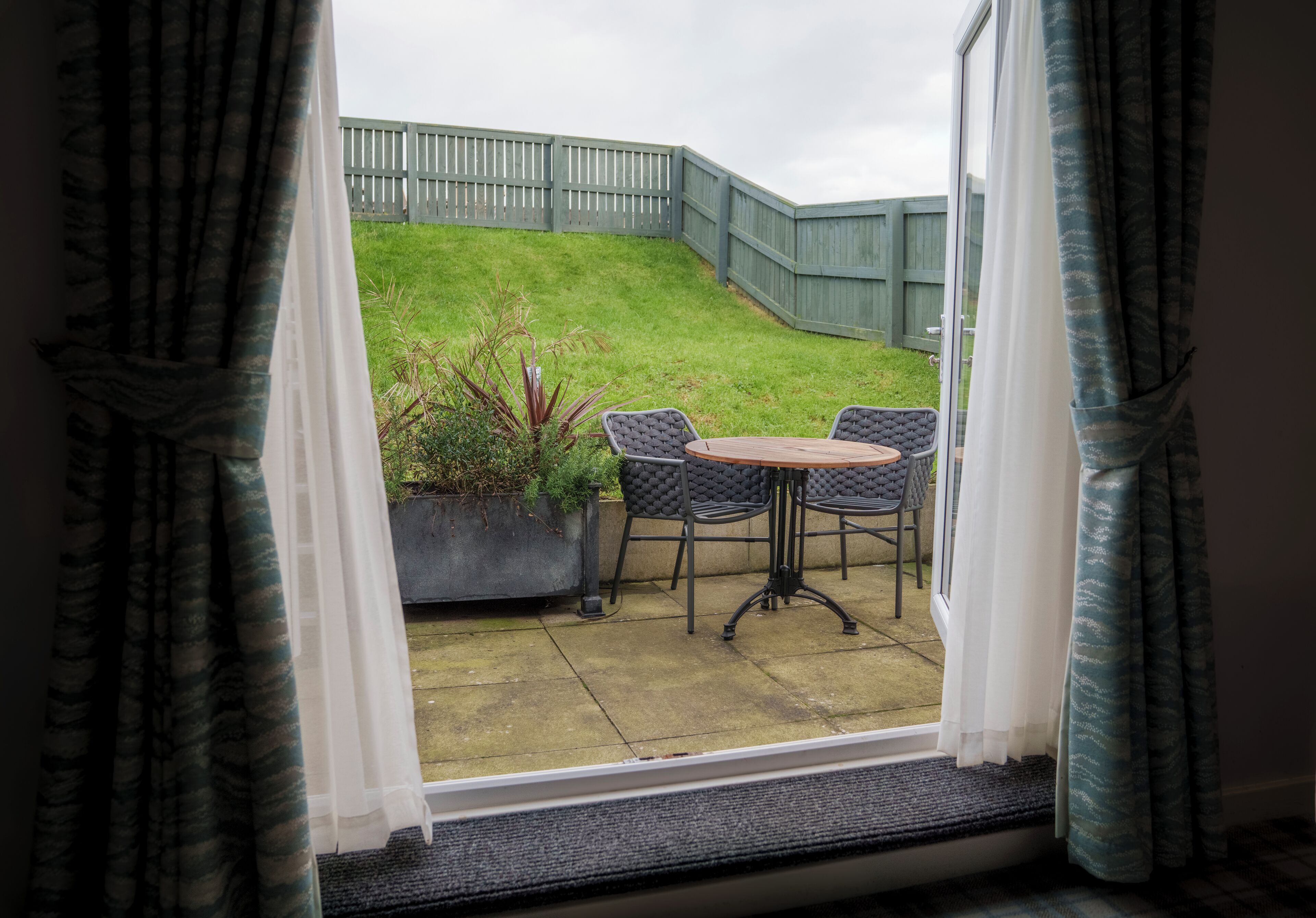 A view from a window to a patio area with a wooden table and chairs, a grassy slope, and a wooden fence.