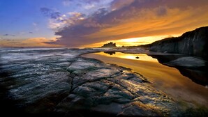 A coastal landscape with a rocky shoreline, calm water reflecting the sky, and a distant castle-like structure.