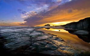 A coastal landscape with a rocky shoreline, calm water reflecting the sky, and a distant castle-like structure.