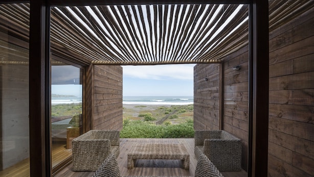 A wooden deck with a woven bench and two side chairs, overlooking a beach. - Playa Lobos Casas & Clubhouse (Pichilemu)