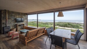 A spacious room with a large wooden table, comfortable seating, a stone wall, a television, and a view of the ocean. - Playa Lobos Casas & Clubhouse (Pichilemu)
