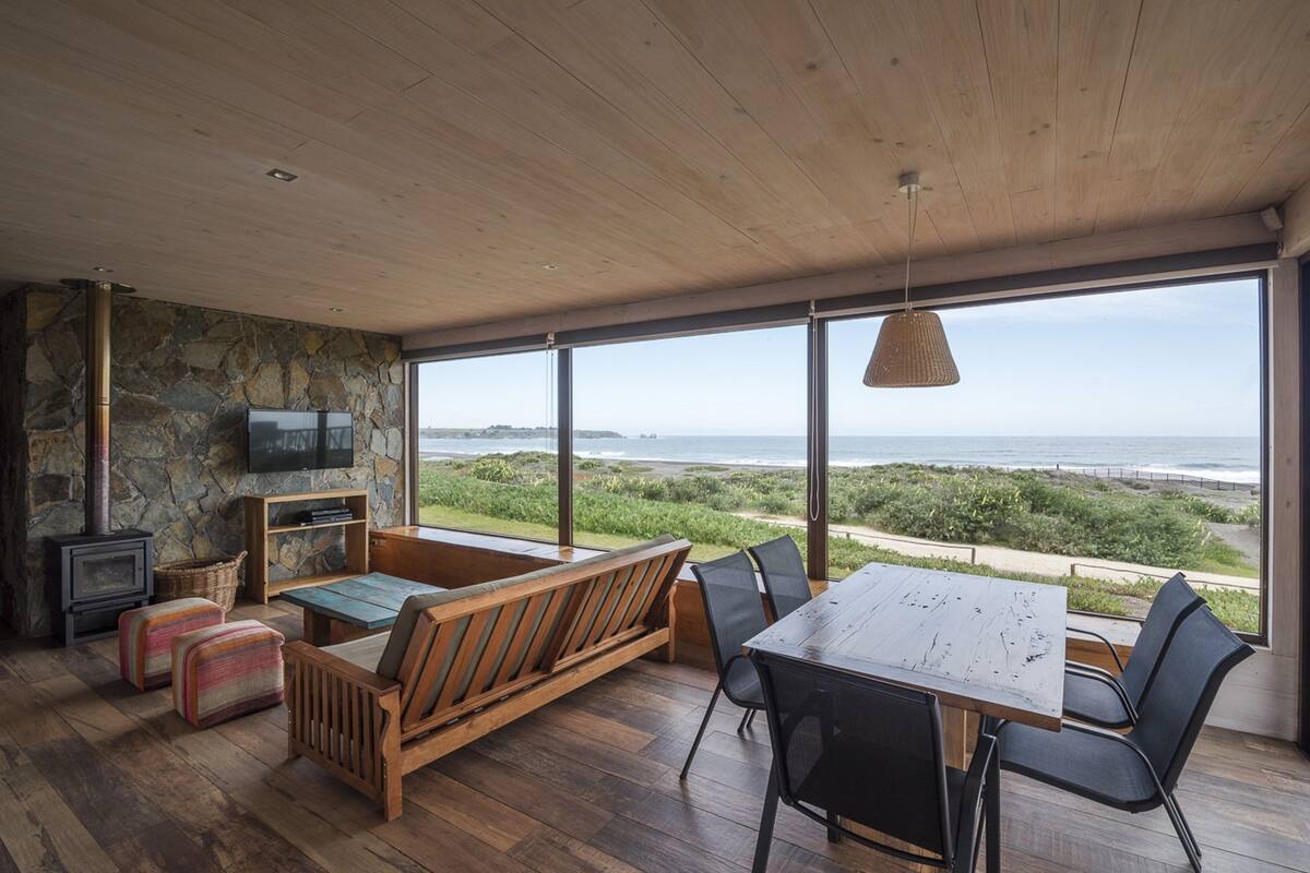 Ein geräumiges Zimmer mit großem Holztisch, bequemen Sitzgelegenheiten, einer Steinwand, einem Fernseher und Blick auf den Ozean.