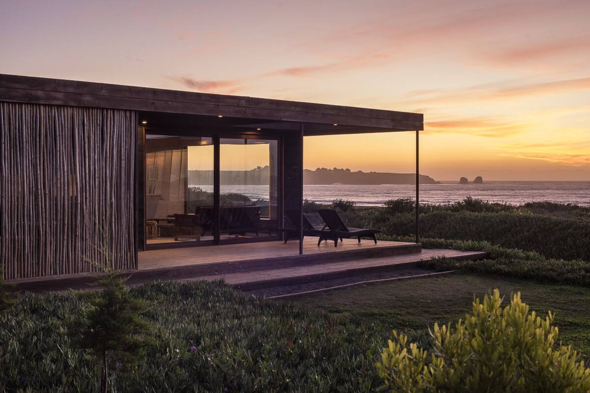 Ein modernes Haus mit Glasfassade, Sitzgelegenheiten im Freien und Blick auf den Ozean im Sonnenuntergang.