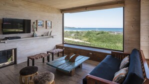 A modern living room with a large window overlooking a beach, wooden flooring, a sofa, a coffee table, and a flat-screen TV mounted on the wall. - Playa Lobos Casas & Clubhouse (Pichilemu)