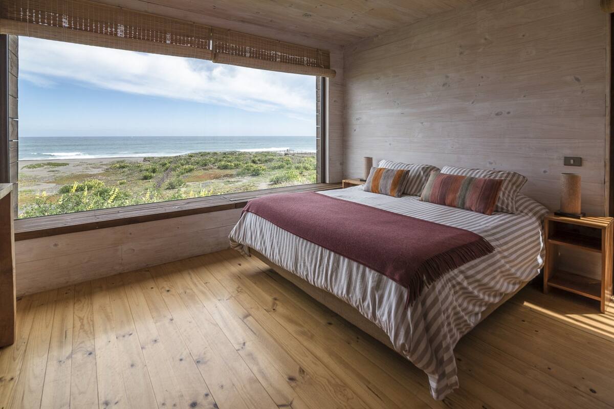 Ein Schlafzimmer mit großem Fenster und Blick auf einen Strand, Holzboden sowie einem Bett mit gestreifter Bettwäsche und Kissen.