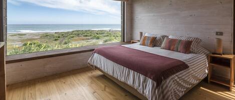 Un dormitorio con una ventana grande que da a una playa, suelo de madera y una cama con ropa de cama y almohadas a rayas.