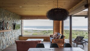 A wooden interior with a large table and chairs, a stone wall, and a view of the ocean. - Playa Lobos Casas & Clubhouse (Pichilemu)