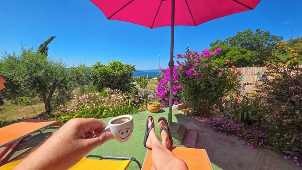 En person som kopplar av under ett rosa parasoll, håller i en mugg med ett leende på och har utsikt över en trädgård och havet.