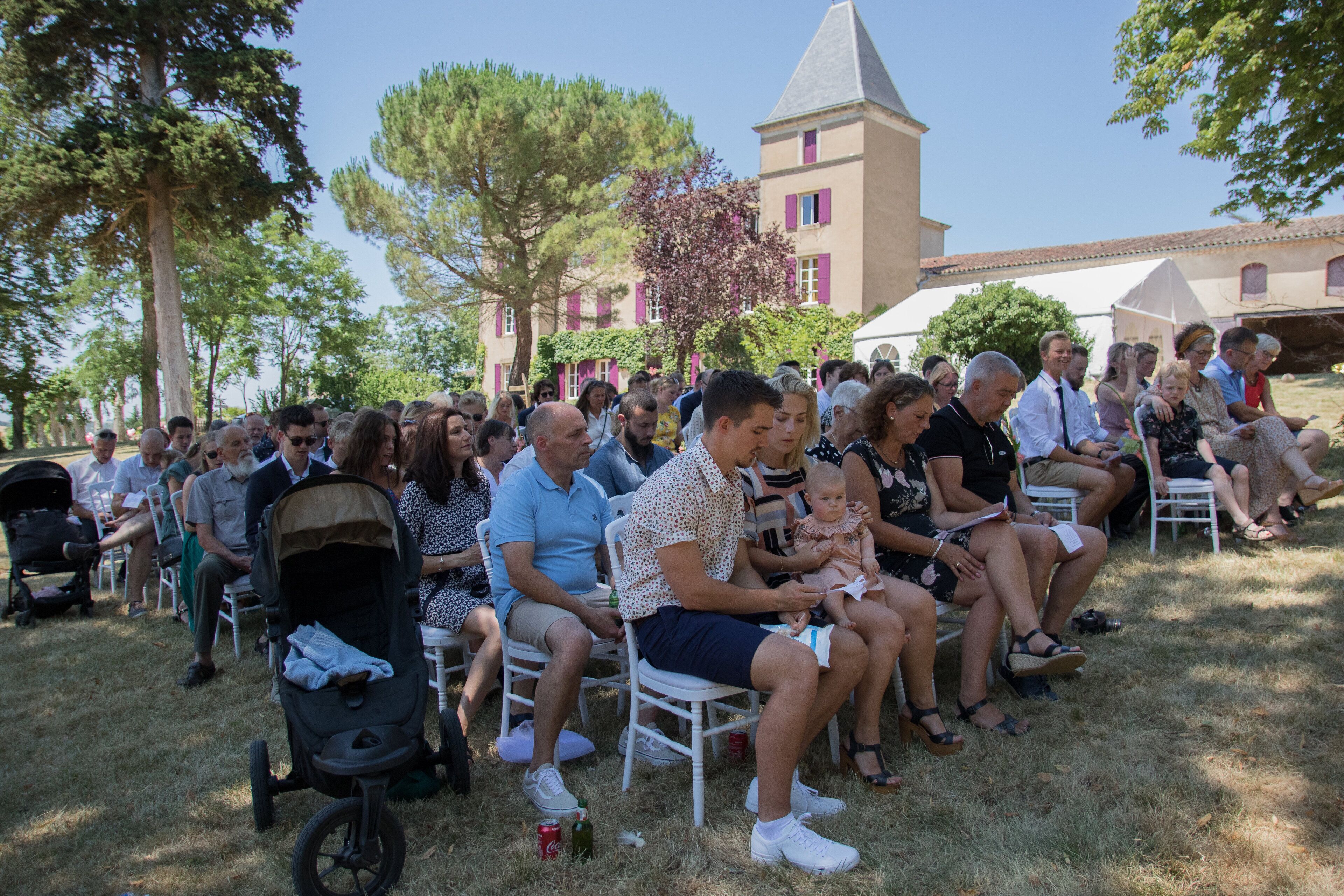 Un groupe de personnes est assis à l’extérieur, sur des chaises, avec un bâtiment en arrière-plan.