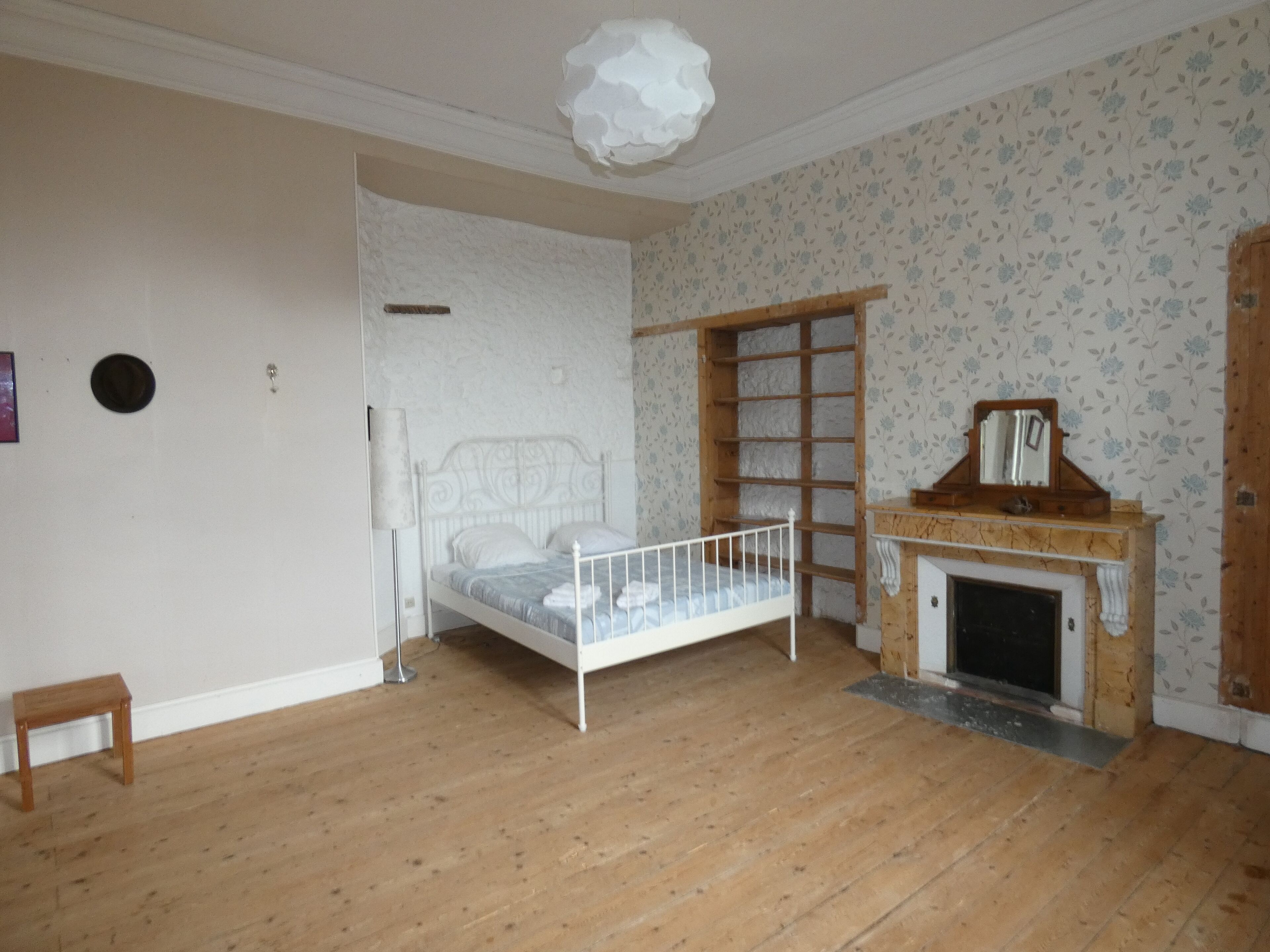 A bedroom with a wooden floor, a white bed, a wooden shelf, and a fireplace.