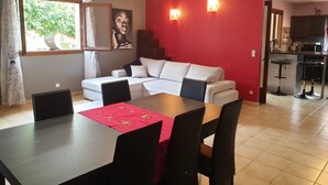 Un coin repas avec une table, des chaises et une nappe rouge. On y trouve un canapé blanc et un mur rouge orné de deux lampes murales. Une fenêtre avec des rideaux est visible, et un espace cuisine se trouve à l’arrière-plan.