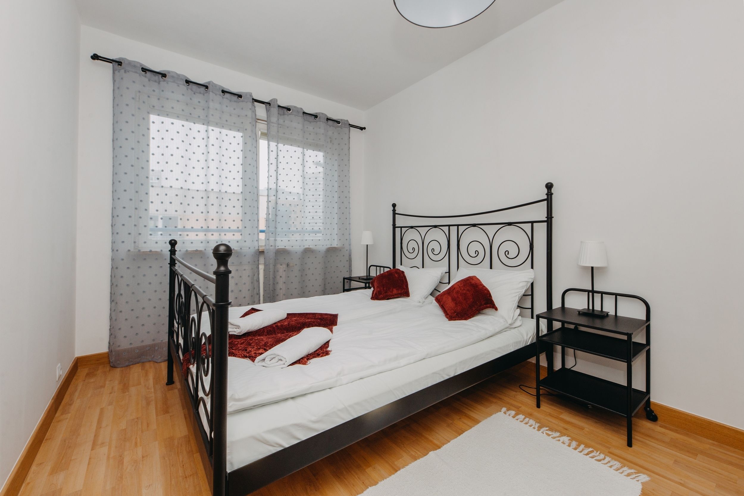 Un lit bien fait, avec du linge de lit blanc et des coussins rouges, une tête de lit en métal noir, une table de chevet avec une lampe et une fenêtre ornée de rideaux légers.