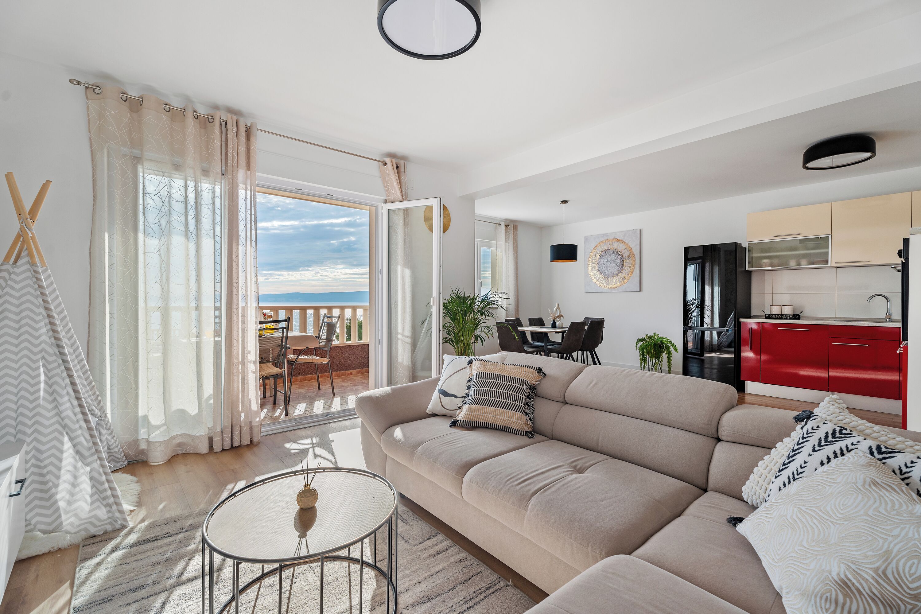 Een moderne woonkamer met een beige bank, een ronde salontafel, en een eetgedeelte met een rode keuken op de achtergrond.