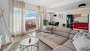 Un salon moderne avec un canapé beige, une table basse ronde et un coin repas, avec une cuisine rouge en arrière-plan.