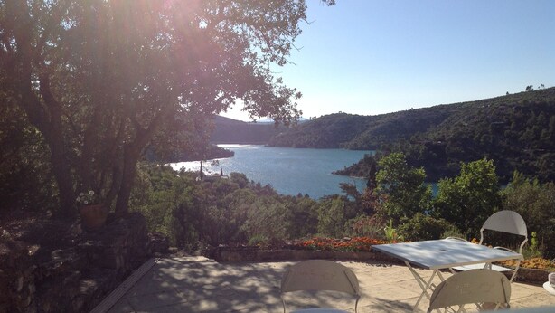 A terrace with a table and chairs overlooking a lake, surrounded by trees and hills.