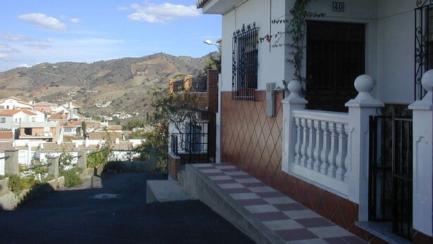 Ein Wohngebäude mit weißer Fassade, einem Balkon mit weißer Brüstung und Blick auf eine hügelige Landschaft im Hintergrund.