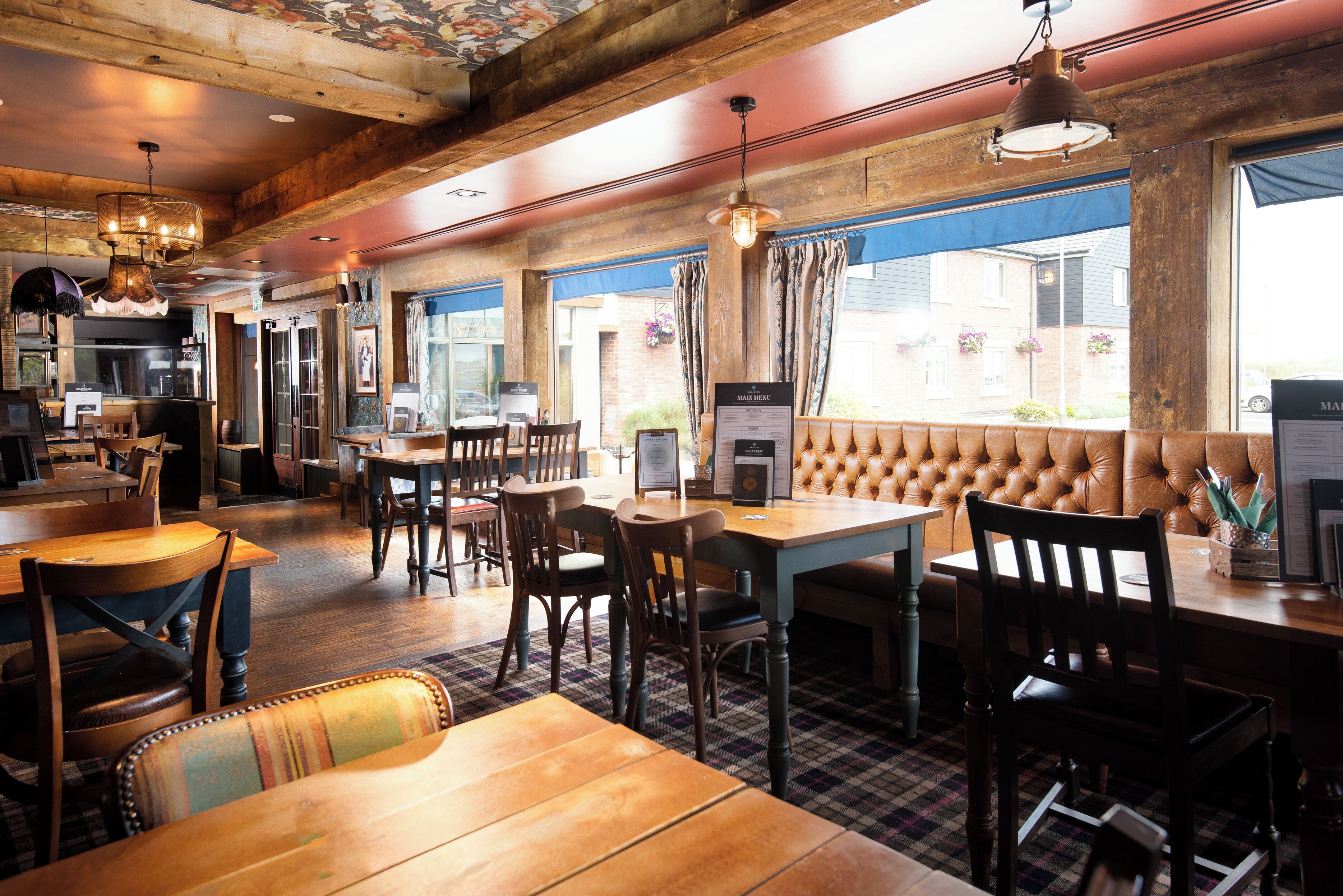 Een restaurantinterieur met houten tafels en stoelen, een bar en grote ramen met gordijnen.