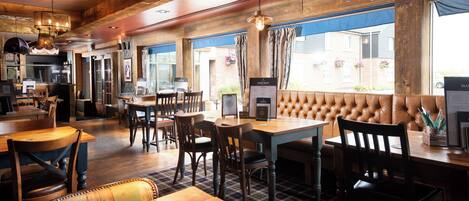 A restaurant interior with wooden tables and chairs, a bar area, and large windows with curtains.