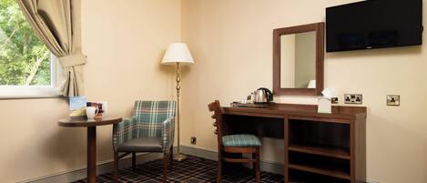 Quarto de hotel com carpete xadrez, escrivaninha com espelho, televisão na parede e janela com cortinas.