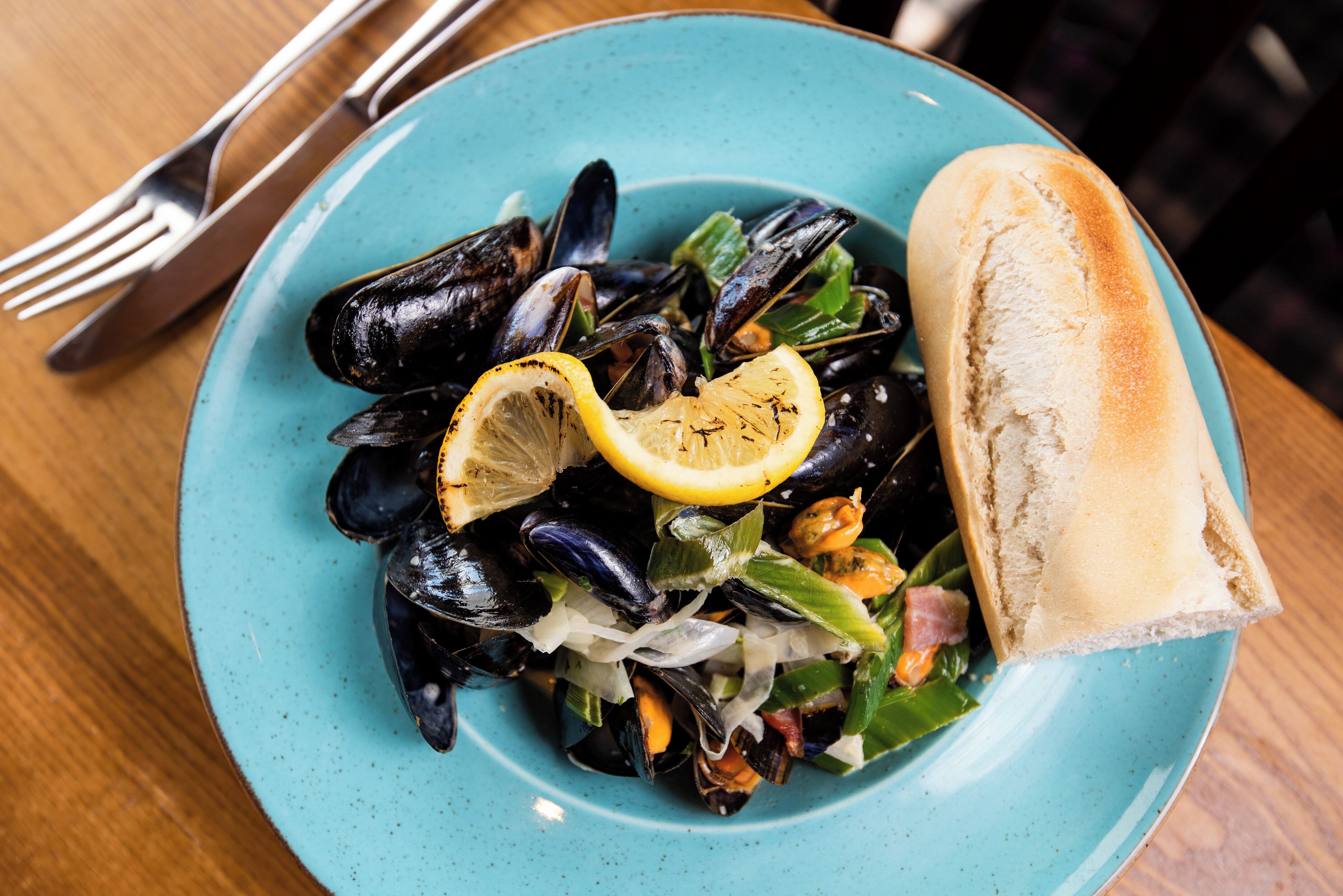 A plate of mussels with lemon and greens, accompanied by a piece of bread on a turquoise plate.