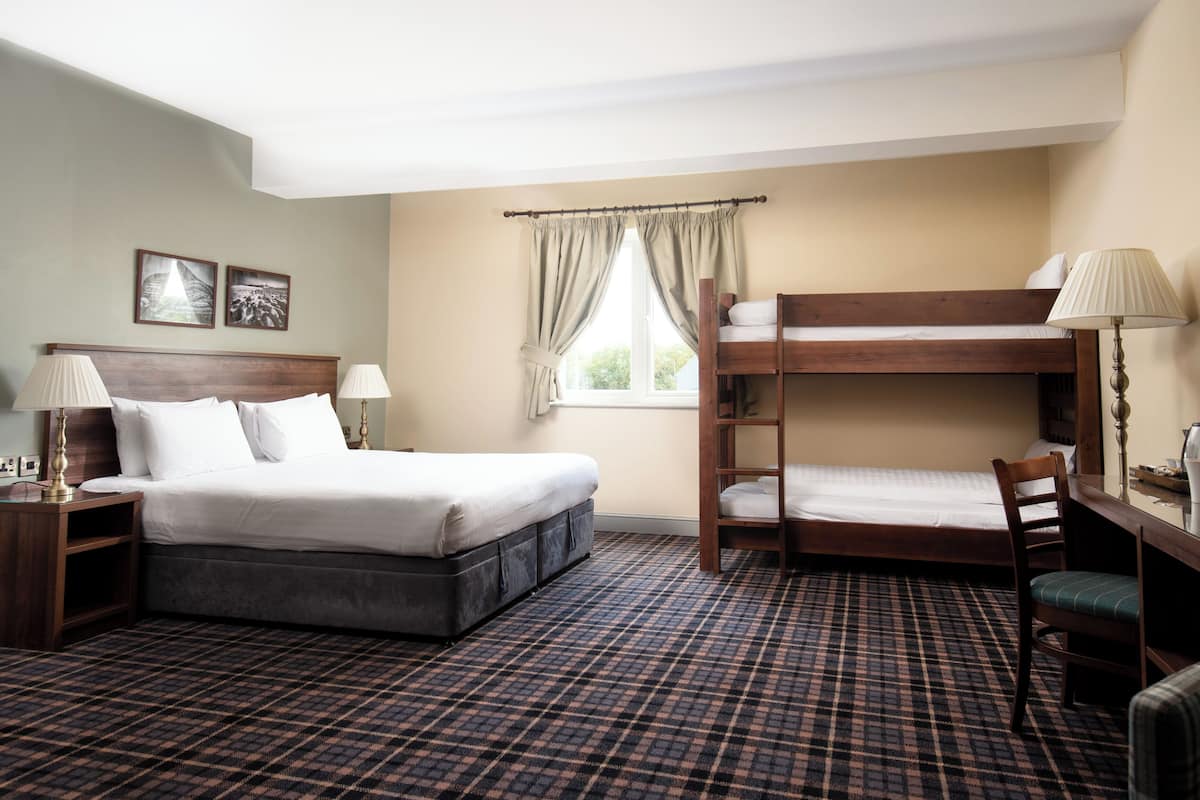 A hotel room with a large bed, a smaller bunk bed, a desk with a chair, and a window with curtains.