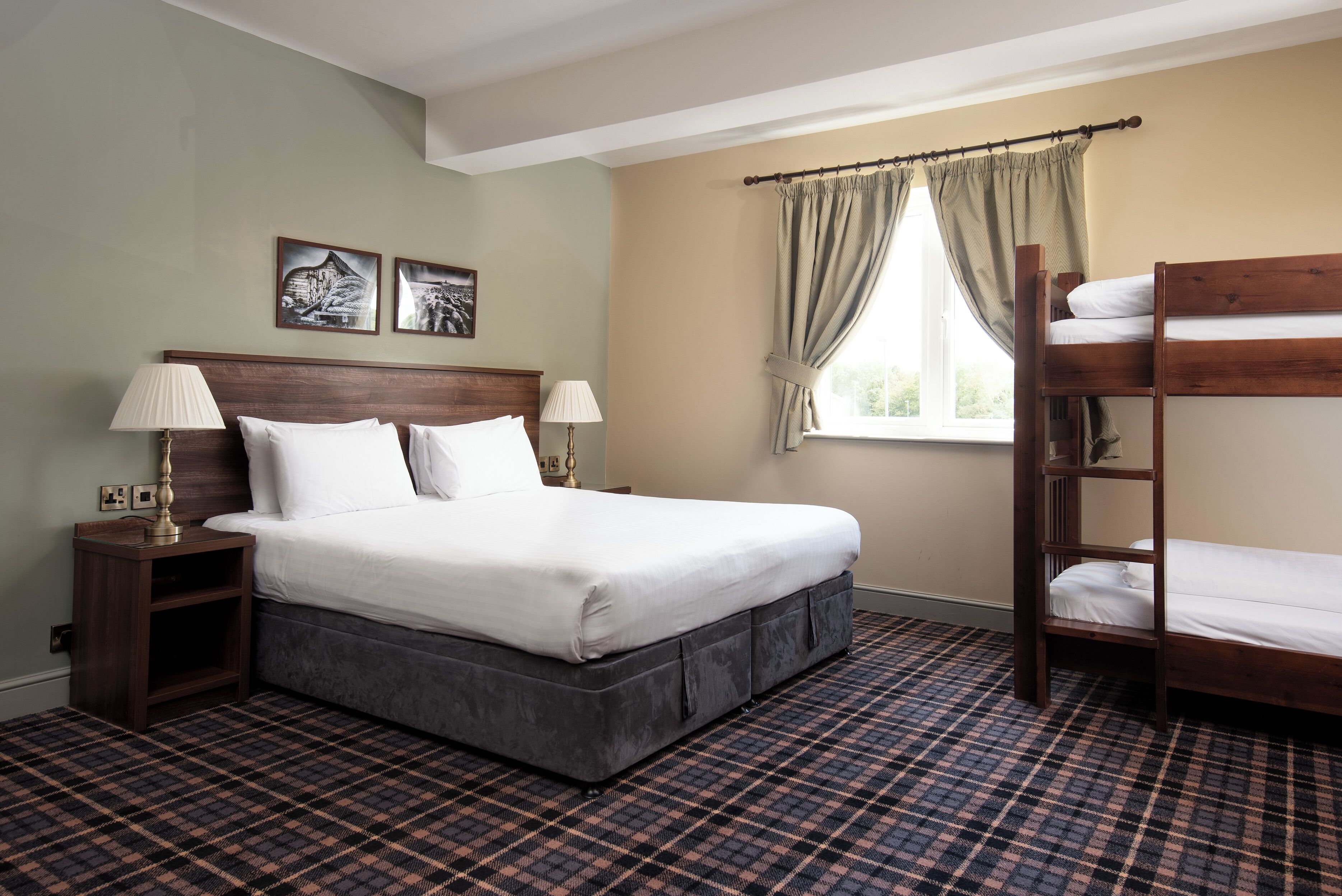 Quarto de hotel com uma cama grande, uma cama beliche de madeira, duas mesas de cabeceira com luminárias, um tapete xadrez e duas fotografias emolduradas na parede.
