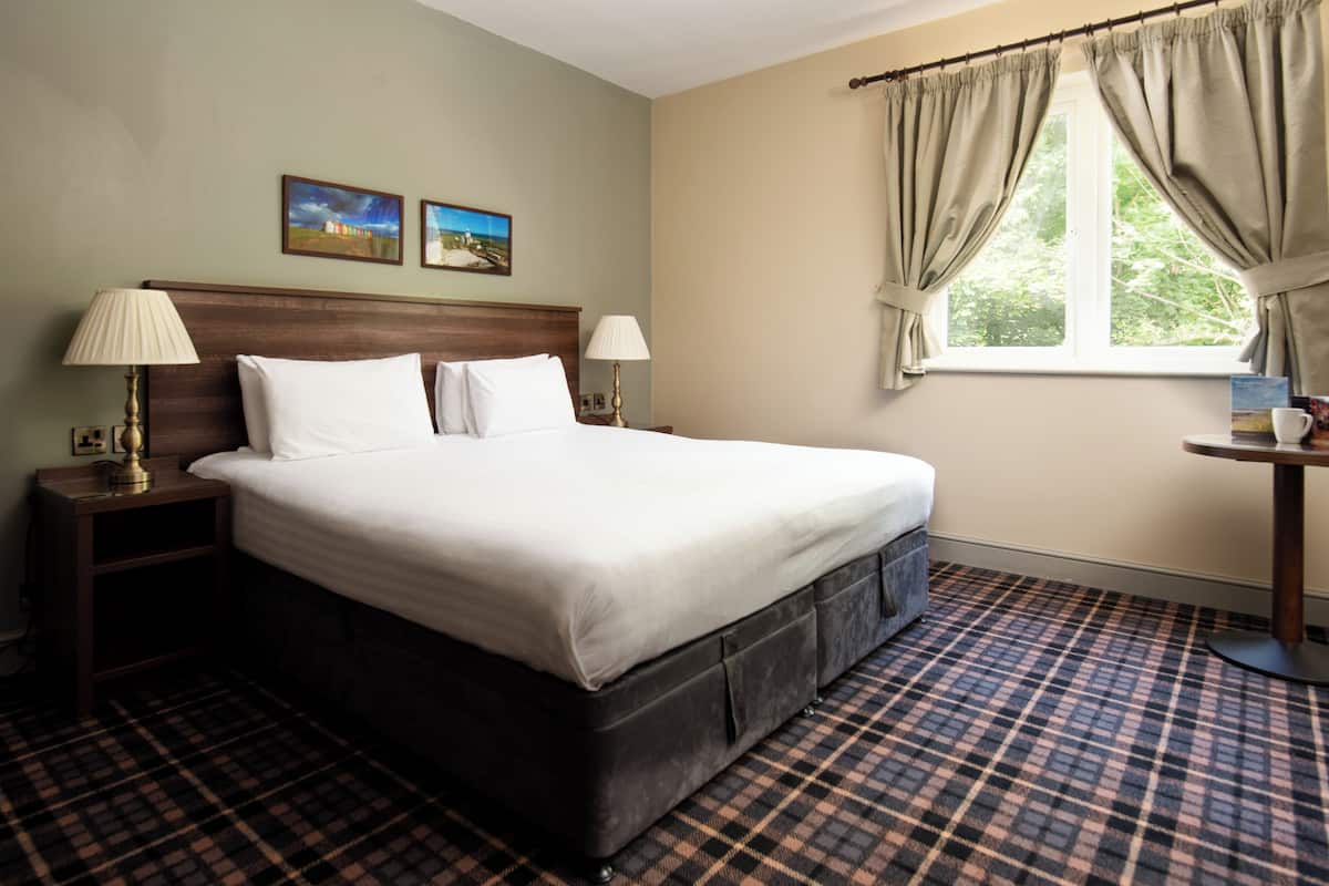 A neatly made bed with a headboard, two bedside lamps, a checkered floor, and a window with curtains.