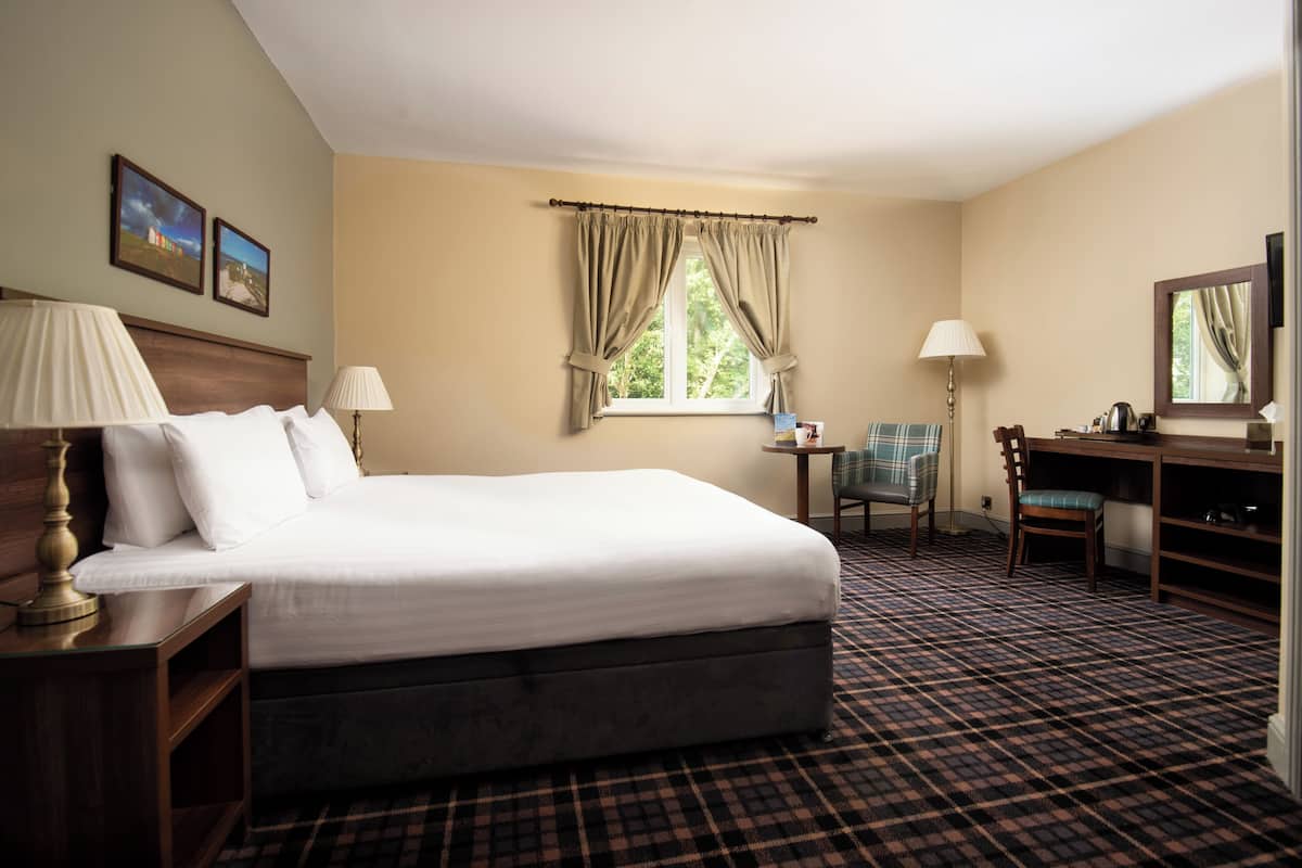 A hotel room with a bed, two bedside lamps, a desk with a chair, a small table, and a window with curtains.