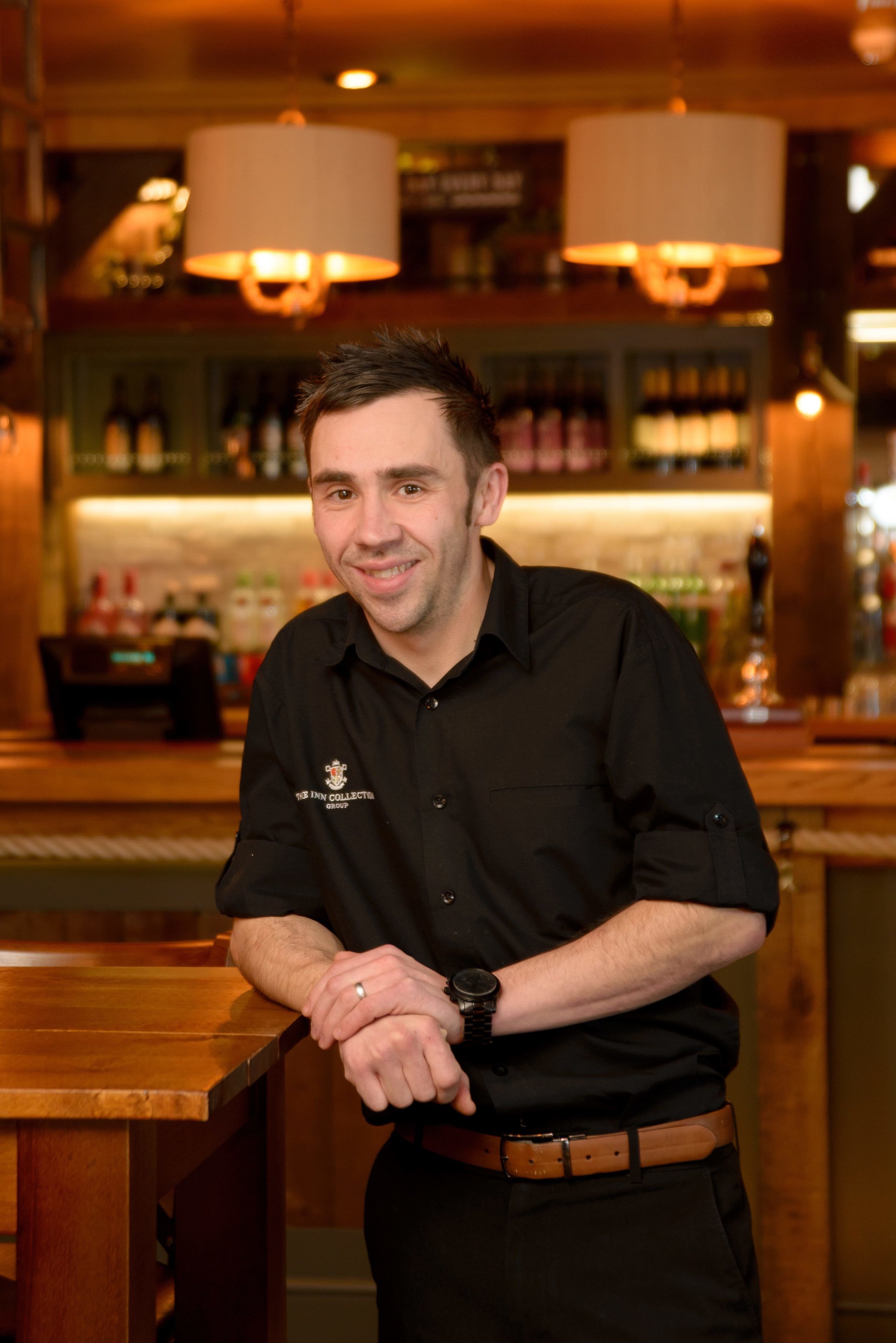 A person standing at a bar with a watch on their wrist, a brown belt, and a ring on their finger.
