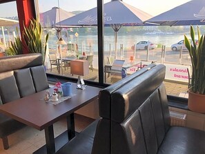 An outdoor seating area with tables and chairs, overlooking a body of water with boats and cars in the background.