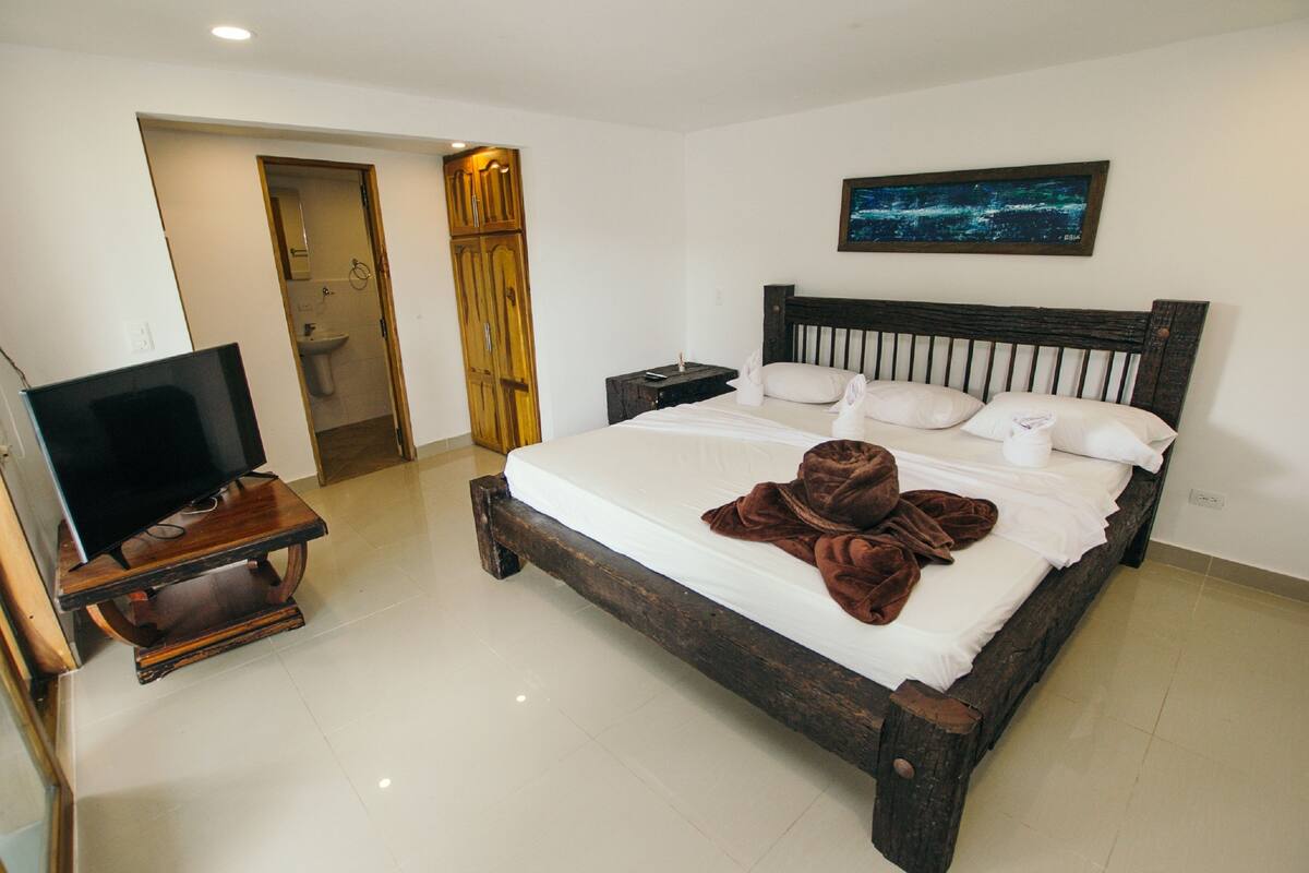 Habitación de hotel con una cama grande, un televisor sobre un soporte de madera, un cuadro en la pared y un baño visible a través de una puerta abierta.