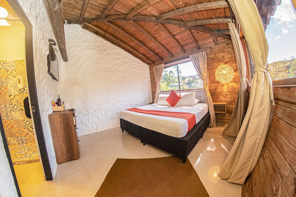 Un dormitorio con techo de madera, una cama con almohada roja, una mesita de noche y una ventana con cortinas.