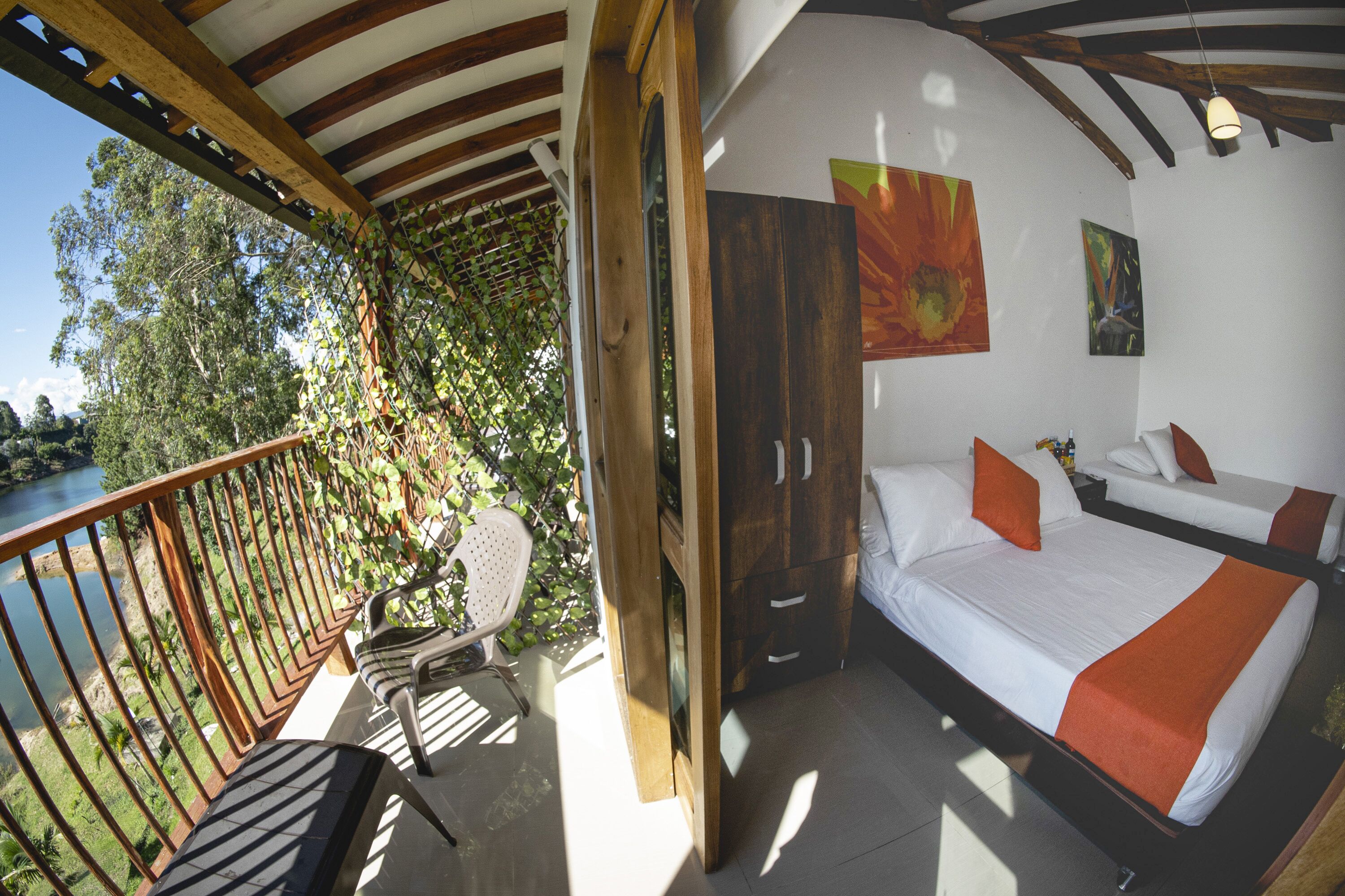A balcony with a view of a river, a bed with white bedding and orange accents, and a wooden ceiling structure.