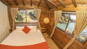 Habitación de cabaña de madera con cama, cortinas, una lámpara colgante y vistas a un lago.