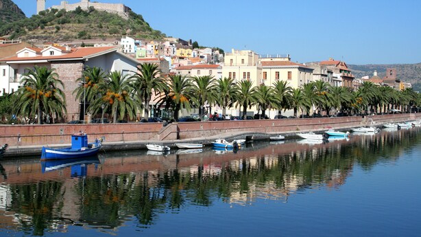 Un poble costaner amb un castell històric sobre una colina, una filera d'edificis colorits i barques amarrades al llarg del front marítim.